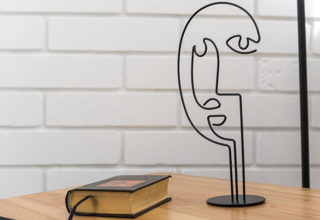 Free A minimalist wire sculpture and book on a wooden table against a white brick wall. Stock Photo