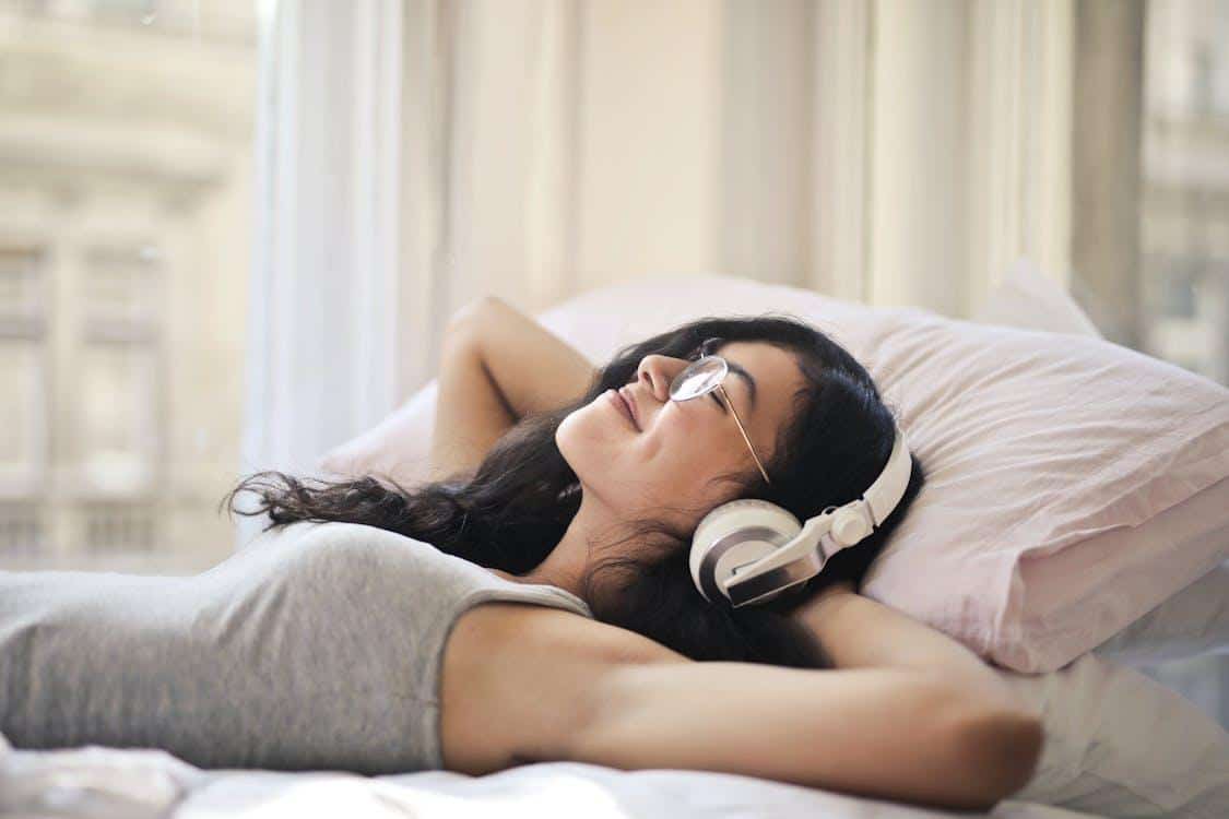 Free A woman with headphones lying on a bed, enjoying music in a relaxed, cozy setting. Stock Photo