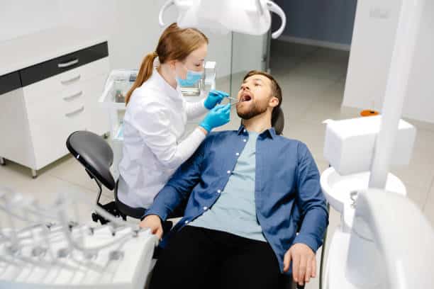 A handsome man is sitting in a chair at a woman's appointment at the dentist's office.
